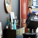 A man moving to a new house carries a large black plastic storage box through the front door. The entryway features a console table with framed art, plants, and bottles, while another storage box rests on the floor.
