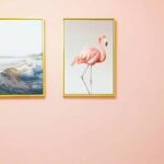 Two framed pictures hang on a pale pink wall—perfect inspiration to choose the right paint color. One frame shows an ocean wave, the other a standing pink flamingo, both with gold edges. The wall is otherwise empty.
